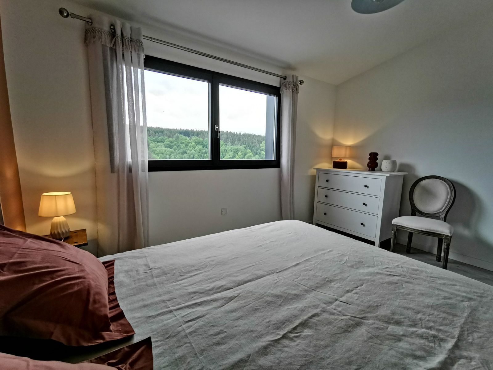 A bedroom with a bed, a nightstand with a lamp, a dresser, a chair, and a window with curtains.