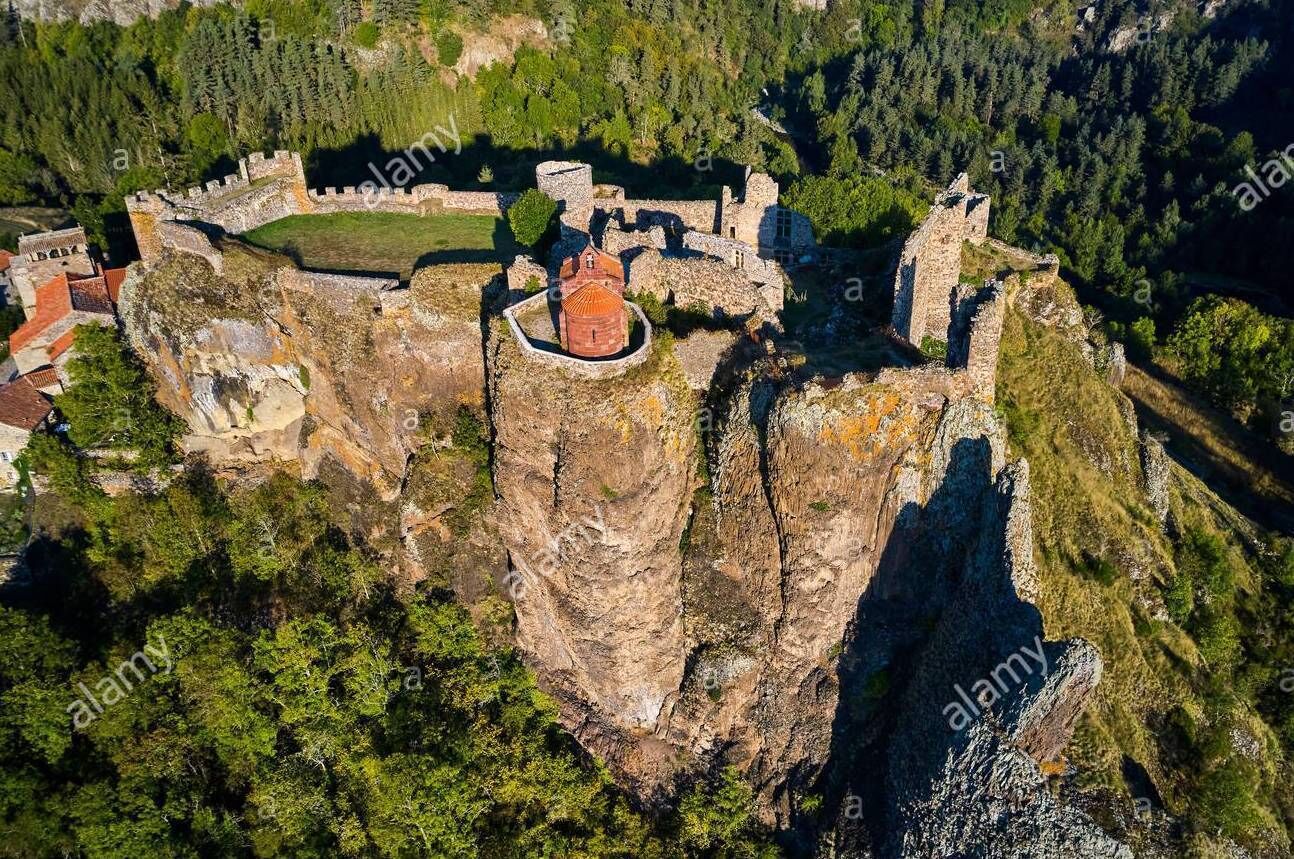 Une vue aérienne d’un château historique perché au sommet d’une falaise, entouré d’une forêt dense.
