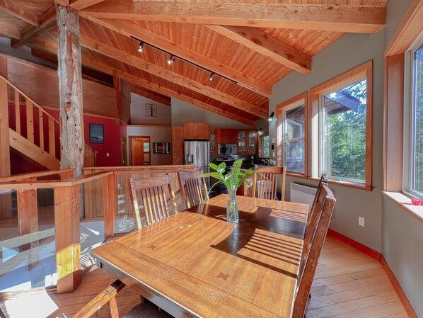 A spacious dining area with a wooden table and chairs, a vase with greenery, and a view of the kitchen and outside through large windows.