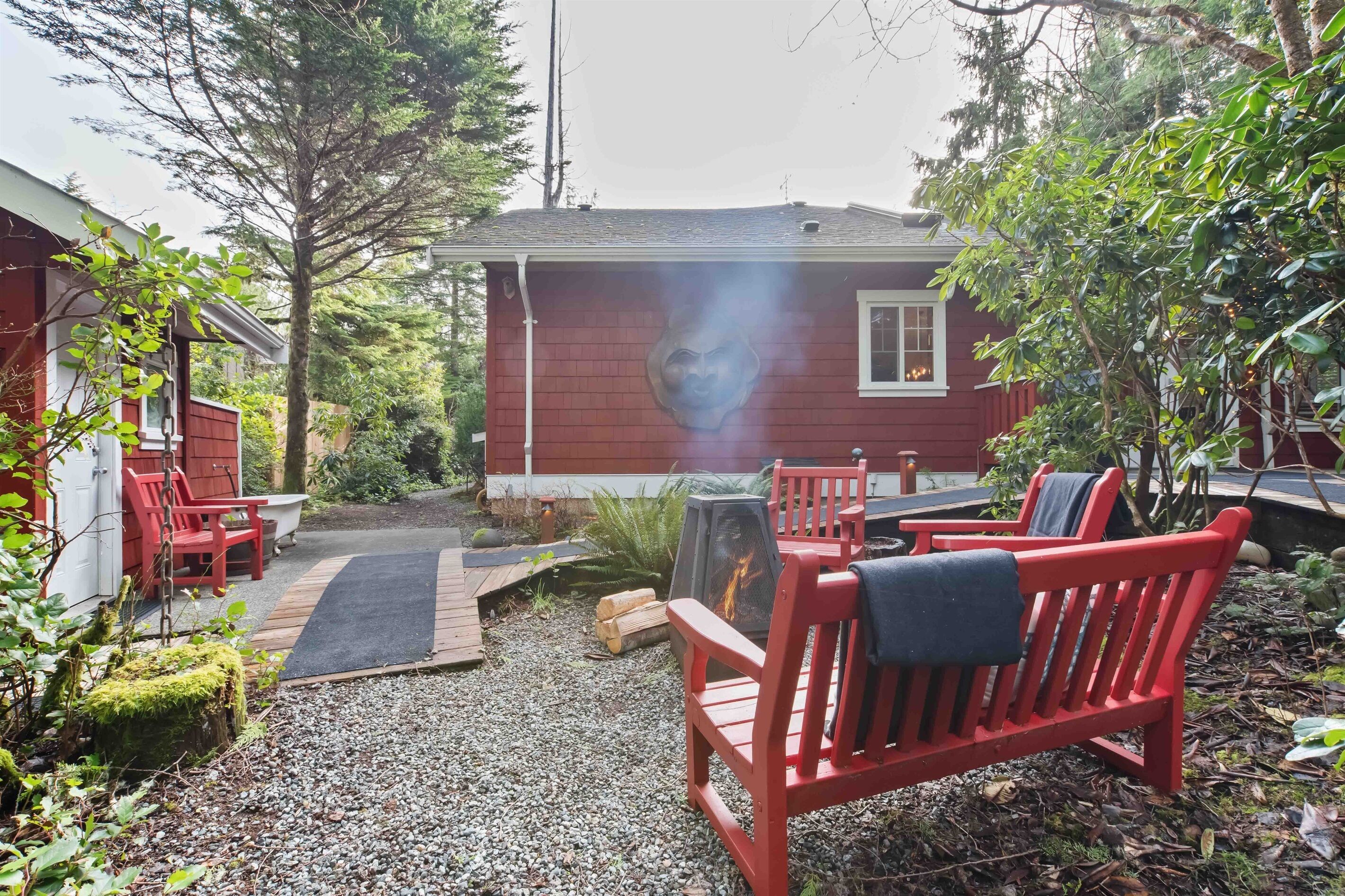 A red cabin with a chimney emitting smoke, surrounded by a gravel path, red chairs, and a small garden area.