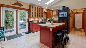 A modern kitchen with wooden cabinets, a central island with stools, a built-in oven, and a large window overlooking greenery. - Coastal Haven-N Chesterman Beach, Hot Tub, Surf! (Tofino)