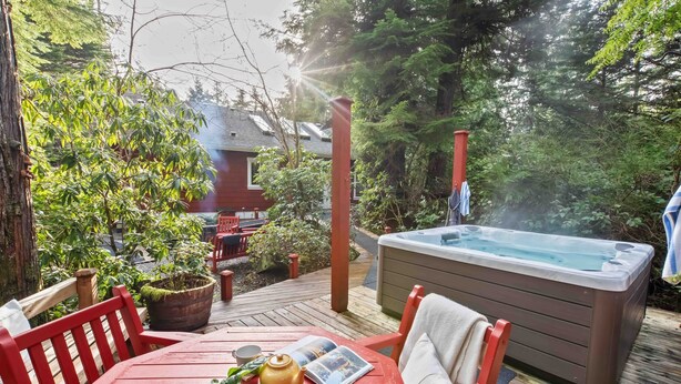 A hot tub on a wooden deck surrounded by greenery, with a red table and chairs nearby. - Coastal Haven-N Chesterman Beach, Hot Tub, Surf! (Tofino)