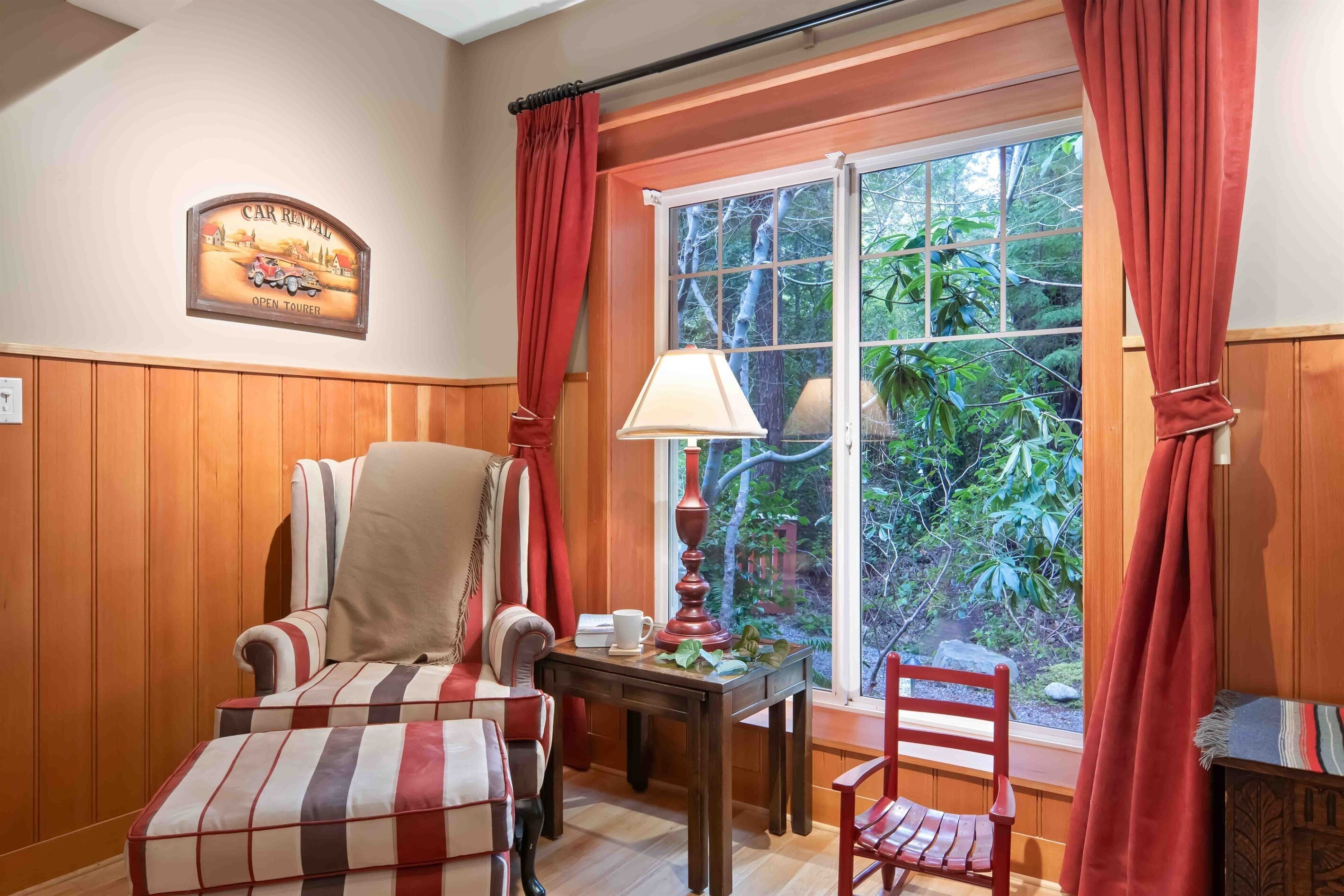 A cozy room with a wooden paneled wall, a striped armchair, a small table with a lamp, and a window with red curtains.