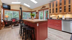 A modern kitchen with a central island, wooden cabinets, and a large window overlooking greenery. - Coastal Haven-N Chesterman Beach, Hot Tub, Surf! (Tofino)