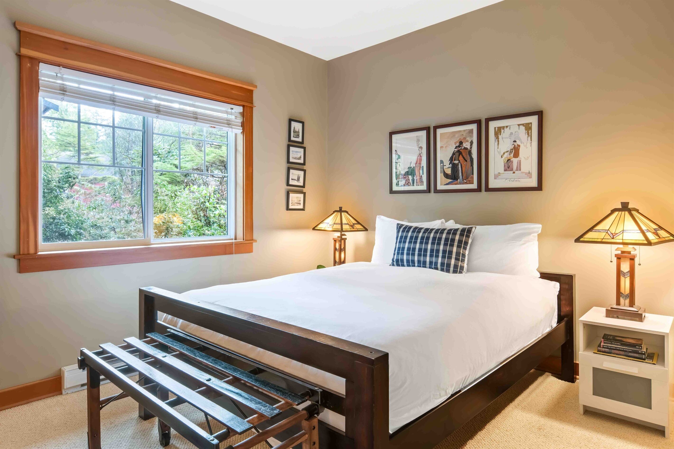 A bedroom with a large bed, two bedside lamps, a bench, a window with blinds, and framed pictures on the wall.
