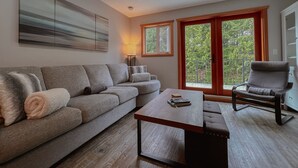 A modern living room with a grey sofa, a wooden coffee table, and a grey armchair. There is a painting on the wall, a lamp, and a view of greenery through the windows.