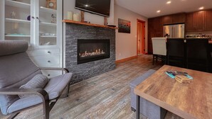A modern living room with a fireplace, a flat-screen TV, a dining area, and a kitchen with wooden cabinets.
