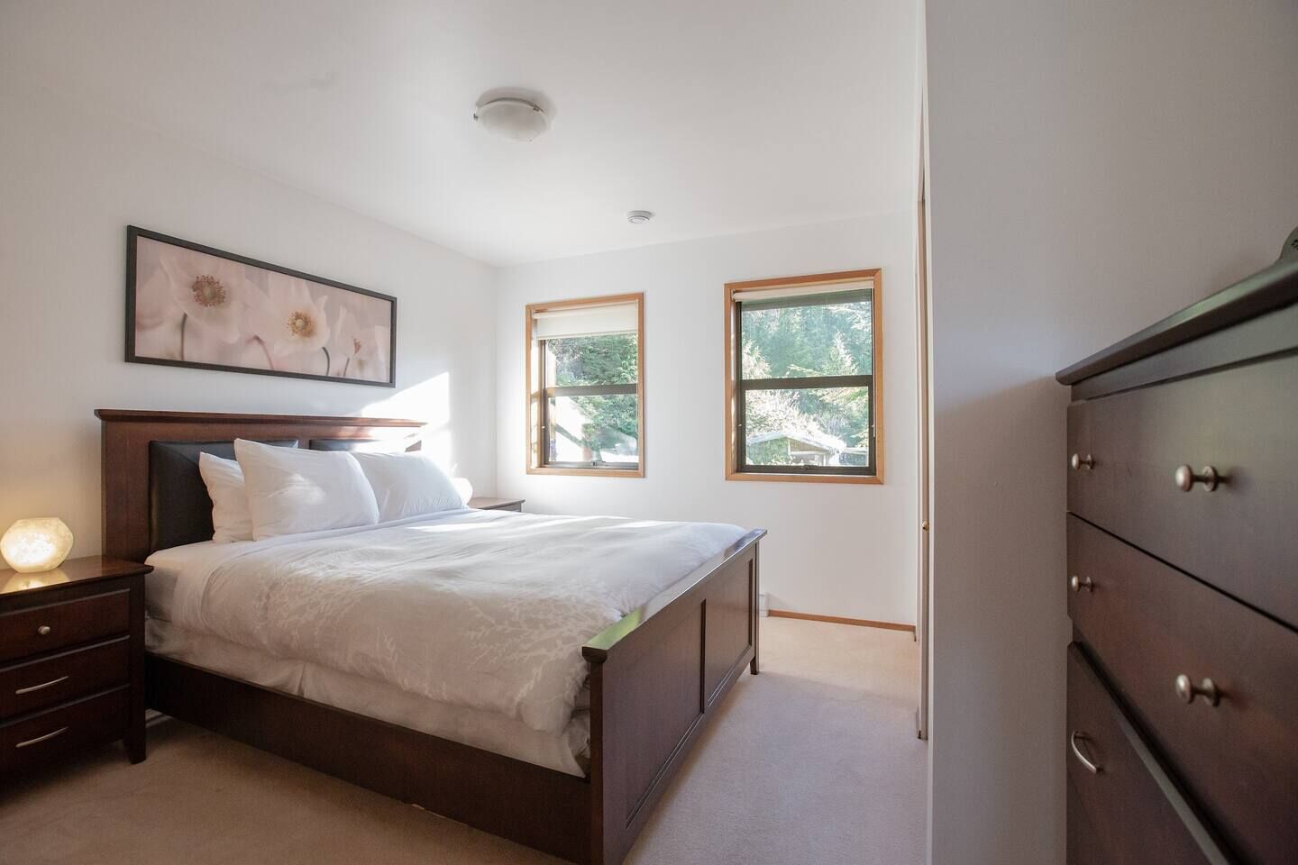 Quarto com cama, mesa de cabeceira, cômoda e duas janelas.