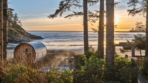 A rustic cabin with a view of the ocean at sunset, surrounded by tall trees and a wooden deck.