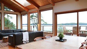 Une pièce spacieuse avec une grande table et des chaises en bois, un canapé en cuir noir et de grandes fenêtres donnant sur un lac et une forêt.