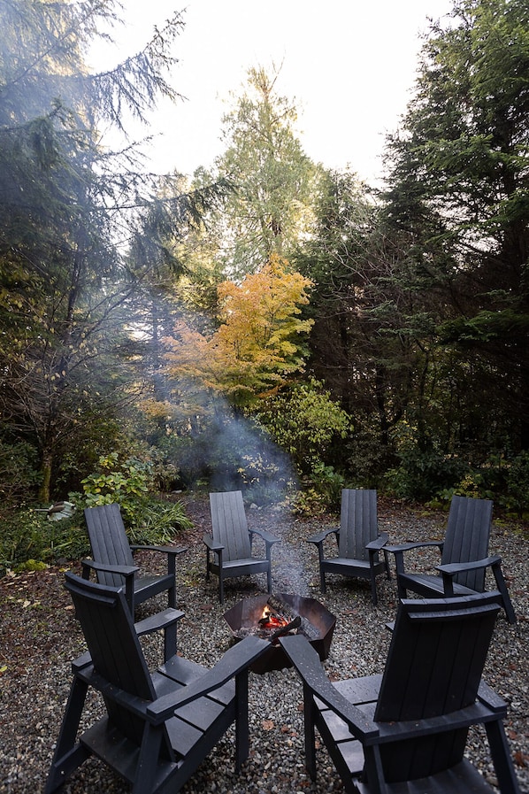 A fire pit with four Adirondack chairs arranged around it, set on a gravel surface with trees in the background.