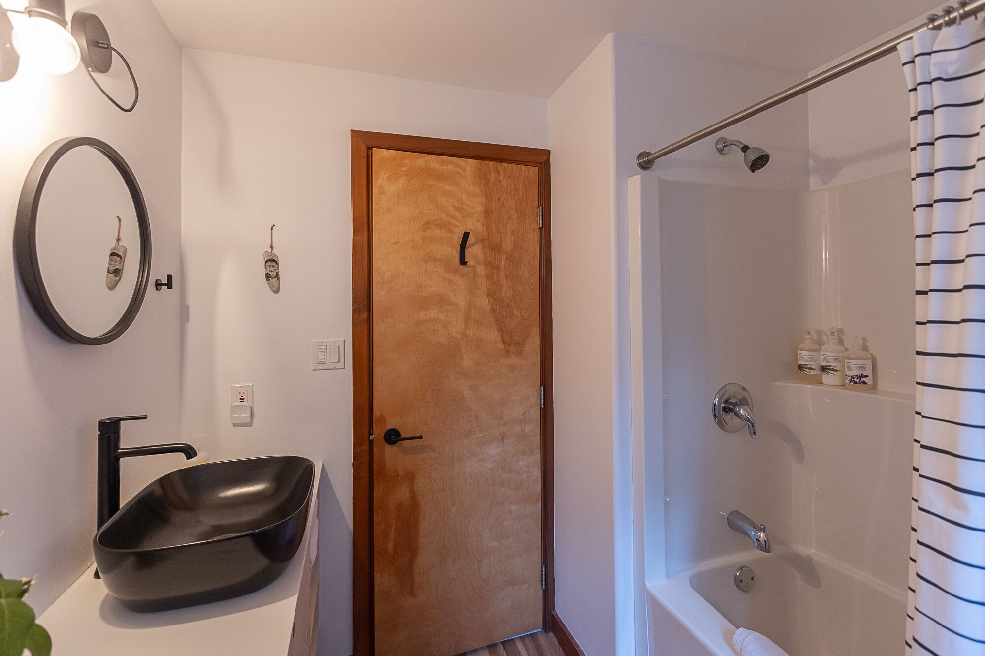 Een badkamer met een houten deur, een zwarte wastafel, een douche met een glazen deur en een handdoekhouder.