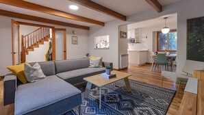 Una sala de estar moderna con un sofá gris, vigas de madera, una escalera, un comedor y una cocina.