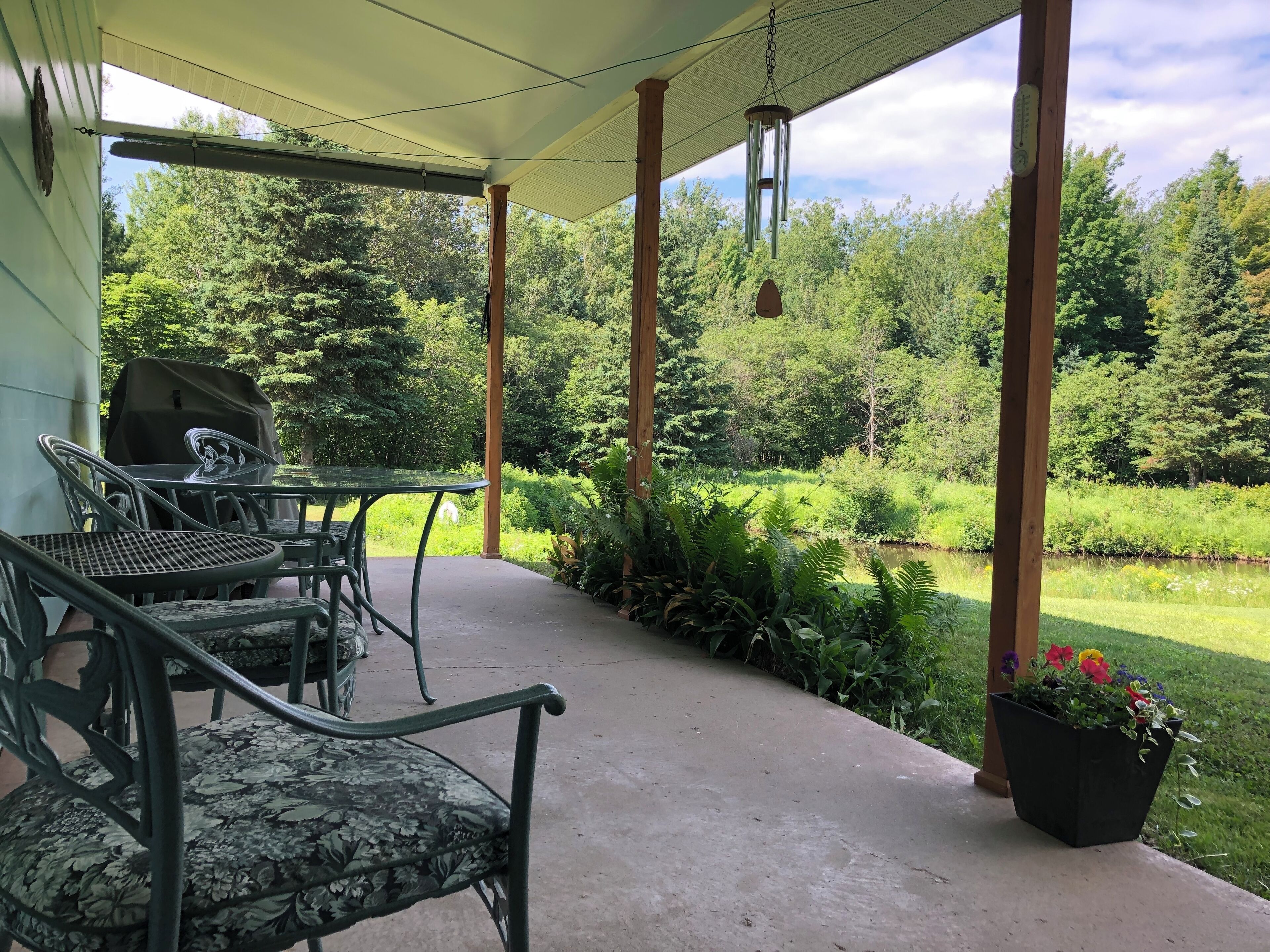 A covered patio with outdoor furniture, a grill, and a view of a lush garden and pond.