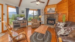 A cozy living room with a stone fireplace, wooden walls, a ceiling fan, and a large window offering a mountain view. - Spectacular Smoky Mtn. Views from Oversized Hot Tub Close to Downtown Gatlinburg (Gatlinburg)