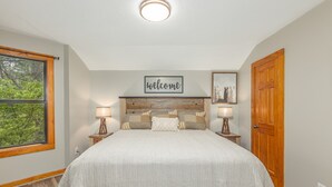 A bedroom with a large bed, a 'welcome' sign above the headboard, two bedside lamps, and a wooden door. - Spectacular Smoky Mtn. Views from Oversized Hot Tub Close to Downtown Gatlinburg (Gatlinburg)