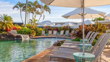 Zona de piscina con sillones de descanso y sombrillas, rodeada de plantas tropicales y palmeras.