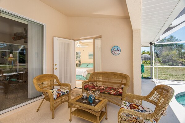Un patio techado con mobiliario de mimbre, una puerta de cristal que da a un dormitorio y vistas a una piscina y follaje.