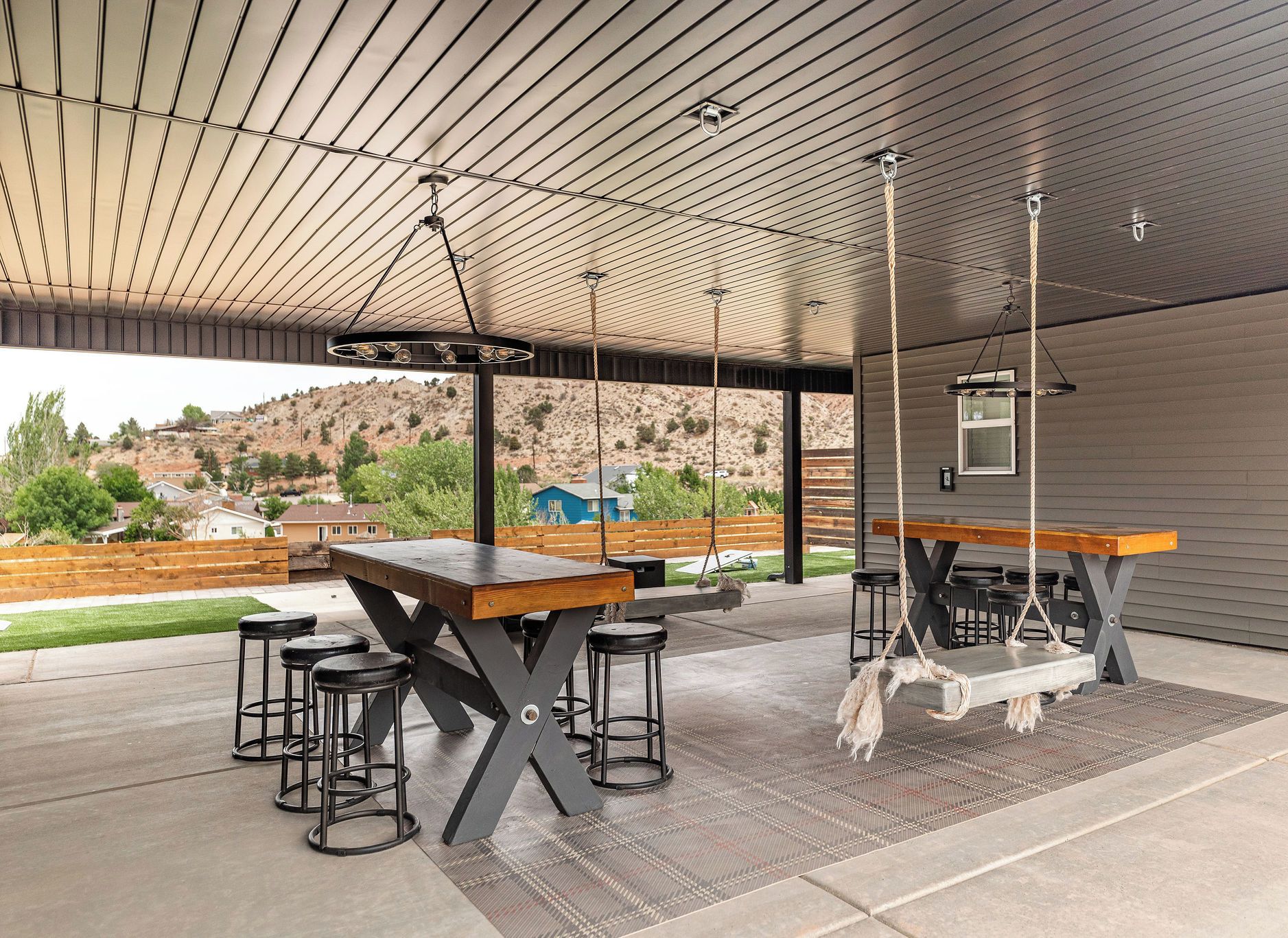 An outdoor patio with a wooden table and bar stools, a hanging swing, and a view of a hilly landscape.