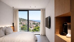 Un dormitorio con una cama grande, televisor, cafetera y vistas a un pueblo a través de un ventanal amplio.