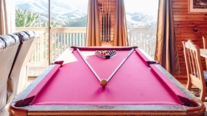 A pool table with a red felt surface, a set of pool cues, and a cue ball. It is located in a room with wooden walls and a view of snow-capped mountains through large windows. - 🏔Sunset Mtn views!⛰ 5 min to PFORGE🚙  HOT TUB! ♨️ (Pigeon Forge)