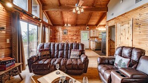 A cozy cabin interior with a leather sofa, a dining table with a basket, and a kitchen area visible through a doorway. - 🏔Sunset Mtn views!⛰ 5 min to PFORGE🚙  HOT TUB! ♨️ (Pigeon Forge)