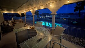 A covered outdoor seating area with a pool, furnished with a glass table and chairs, and string lights.