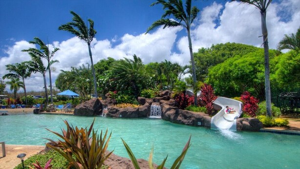 Piscina de un resort tropical con tobogán acuático, rodeada de exuberante vegetación y palmeras.