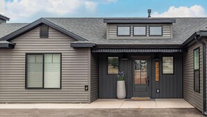Une maison moderne à deux étages, avec un revêtement extérieur gris, une porte d’entrée ornée d’un grand pot de fleurs et un petit porche.