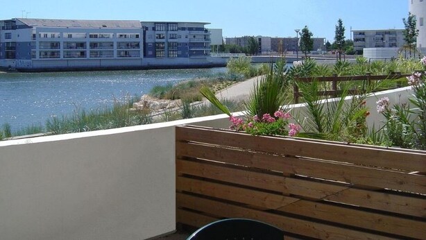 Ein Balkon mit einer Holzbank und einem Blumentopf, mit Blick auf ein Gewässer und moderne Gebäude.