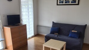 Ein Wohnzimmer mit Holzboden, einer blauen Couch mit Kissen, einem Couchtisch aus Holz, einem Fernseher auf einer Holzkommode und einer Deckenleuchte.