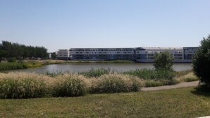 Ein moderner Gebäudekomplex mit mehreren Stockwerken, einem Gewässer im Vordergrund und einer Wiese mit hohem Gras.