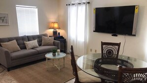 Una sala de estar con un sofá, una mesa de vidrio y dos sillas. Hay un televisor montado en la pared y una lámpara sobre una mesita.