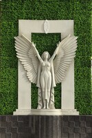 Una estatua de un ángel con alas extendidas, sobre un fondo de seto verde.