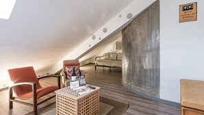 Ein modernes Schlafzimmer im Loft-Stil mit schrägen Wänden, einem großen Fenster, Holzboden, einem roten Sessel, einem beigen Sofa und einem kleinen Tisch mit einer Flasche und einem Willkommensschild.