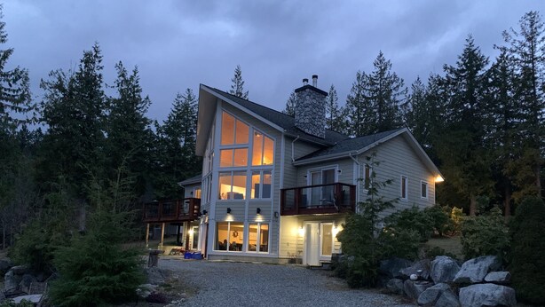 A modern two-story house with a large balcony, a stone chimney, and a gravel driveway surrounded by trees.