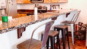 A kitchen with wooden cabinets, a granite countertop, a stainless steel refrigerator, a microwave, and a chandelier. There are three bar stools and a coffee machine.