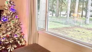 Ein Zimmer mit einem großen Fenster, einem mit Lichtern geschmückten Weihnachtsbaum und Blick auf einen von Bäumen gesäumten, eingezäunten Außenbereich.