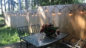 Eine Holzterrasse mit einem Glastisch und Metallstühlen, umgeben von einem Holzzaun und Lichterketten.