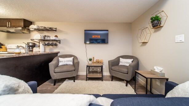 A modern living room with a kitchen area, a TV mounted on the wall, and two armchairs with 'welcome' pillows. - ★Bright Modern Suite ★ Netflix - WiFi ✔Long Stays ★ Clean (Edmonton)