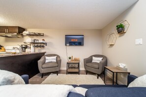 A modern living room with a flat-screen TV mounted on the wall, two grey armchairs with 'welcome' pillows, a small wooden side table with a glass of wine, and a kitchen area with cabinets and a stove.