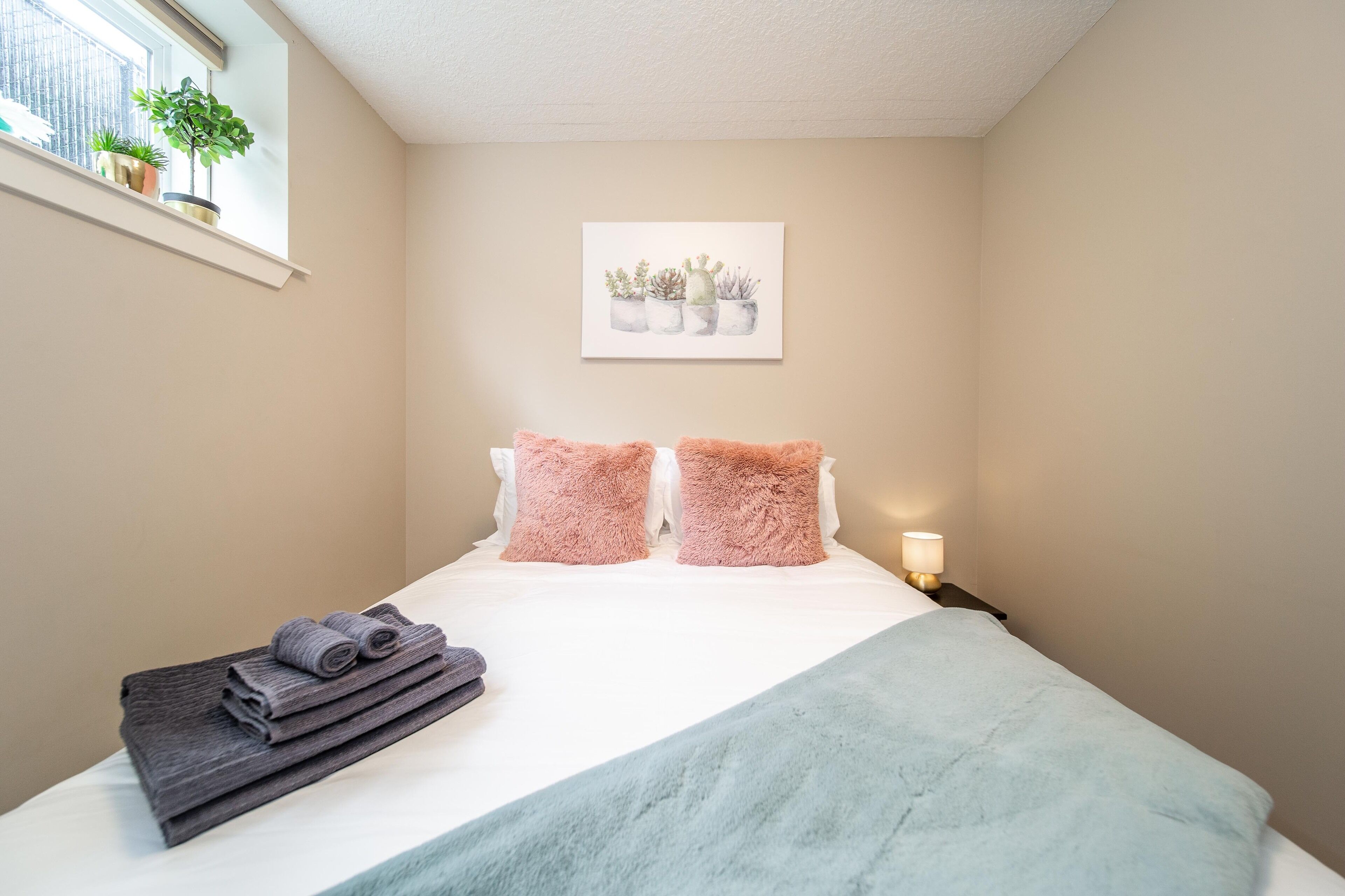 A neatly made bed with a light blue blanket, two pink fluffy pillows, and a stack of folded towels.