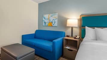 Habitación de hotel con un sofá azul, un pequeño reposapiés, una cama con ropa blanca, una mesita de noche con lámpara, un teléfono y un cuadro en la pared.