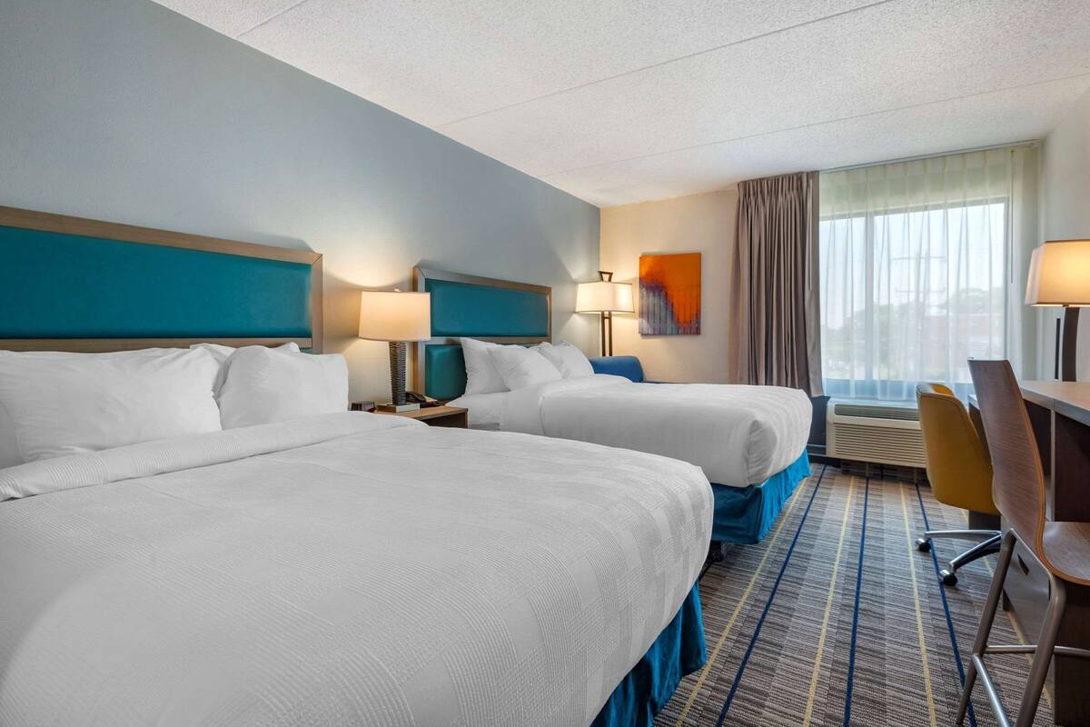 A hotel room with two beds, a desk, a chair, and a large window.