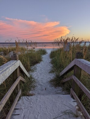 A sandy beach path with wooden railings leading to the ocean, a sign indicating beach access, and a sunset in the background.
