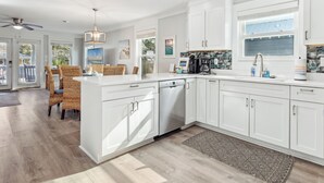 Una cucina moderna con mobili bianchi, lavastoviglie e macchina per il caffè. C'è un'area pranzo con sedie in vimini e una ventola a soffitto.
