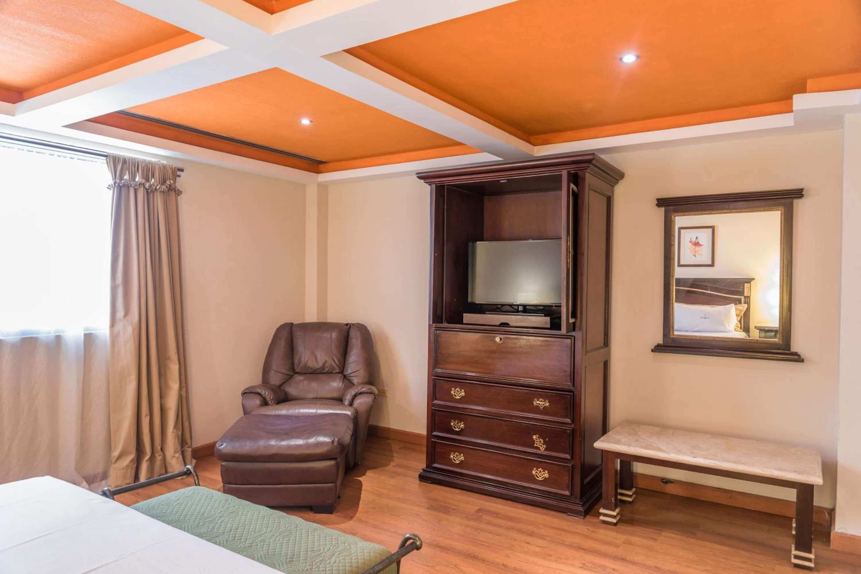 Una habitación con techo de madera, un sillón de cuero, un televisor sobre un soporte de madera, un banco y un espejo fijado a la pared.