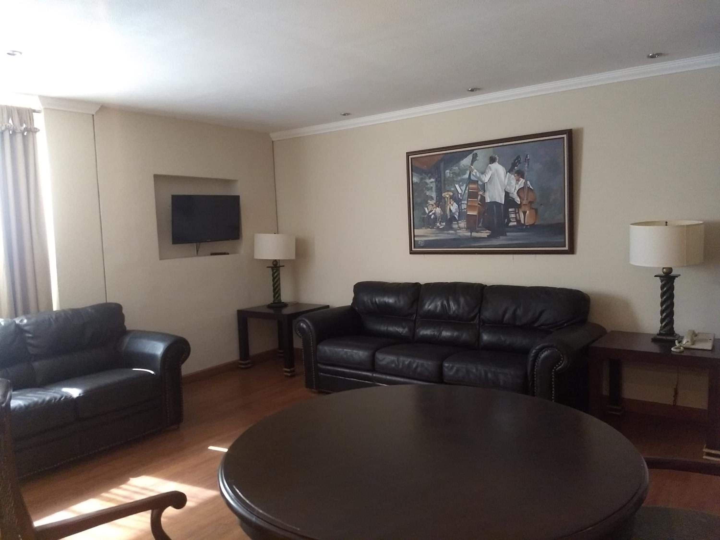 A living room with a black leather sofa, a round coffee table, a flat-screen TV mounted on the wall, and a painting above the sofa.