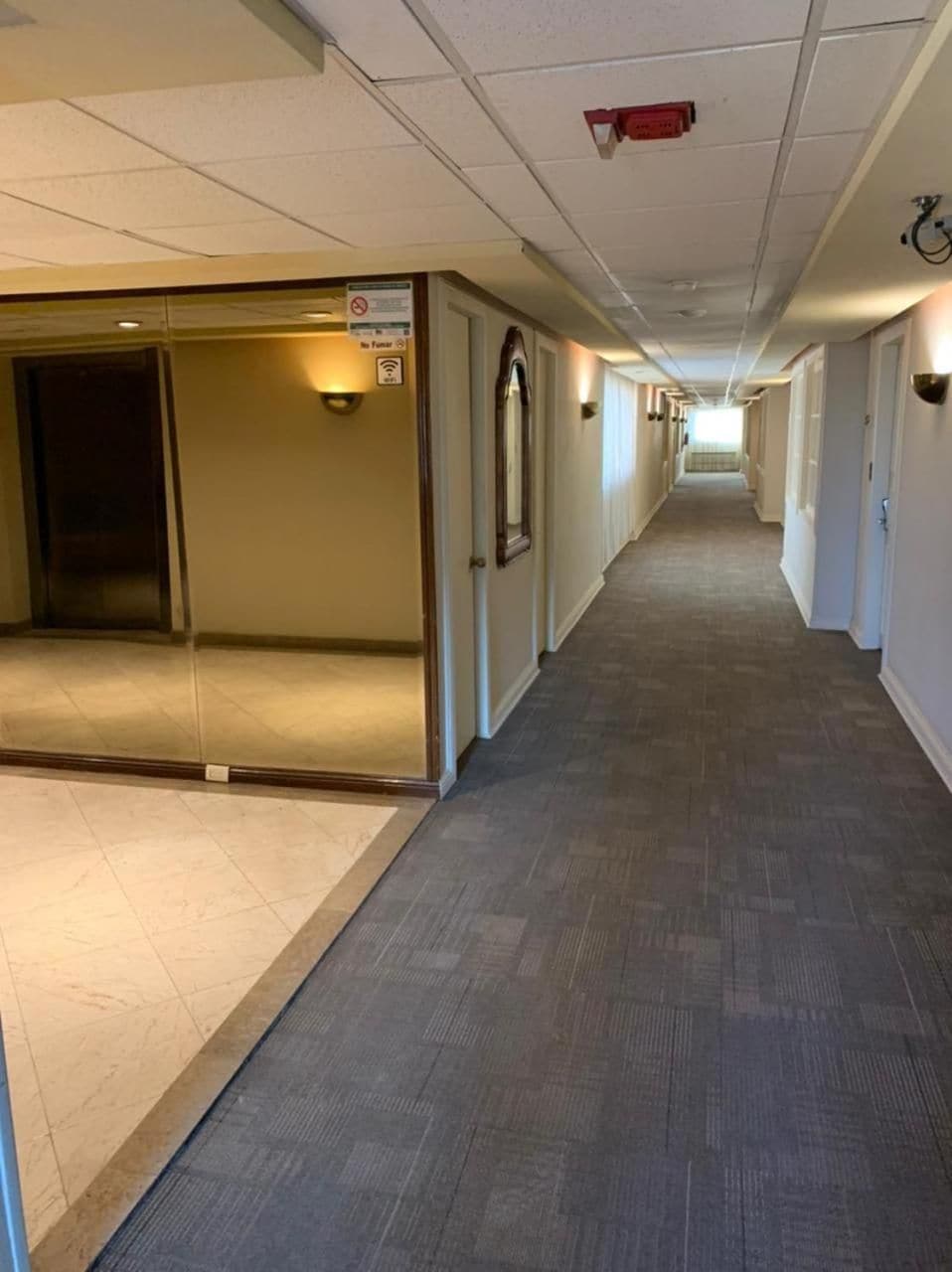 A long, narrow hallway with a tiled floor, a fire alarm on the ceiling, and a door at the end.