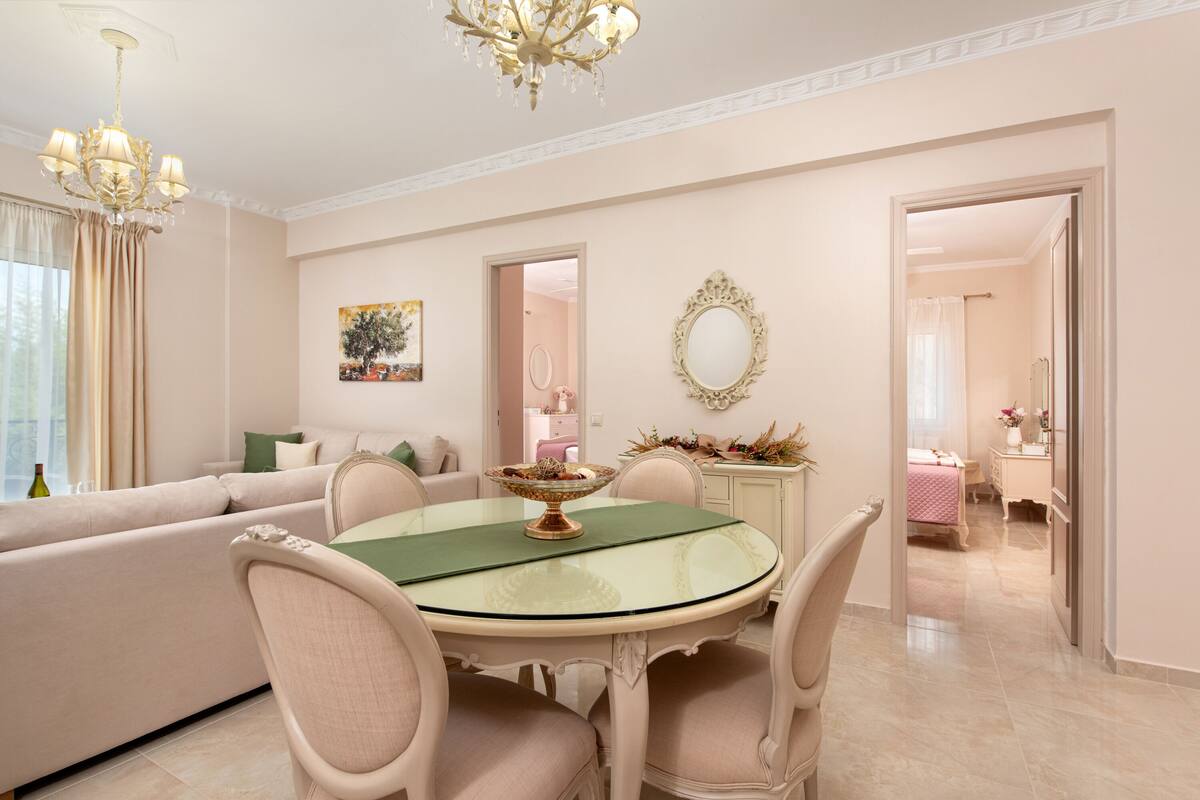 Un comedor con una mesa redonda de vidrio, rodeado de sillas, en una habitación con paredes color crema y una araña.